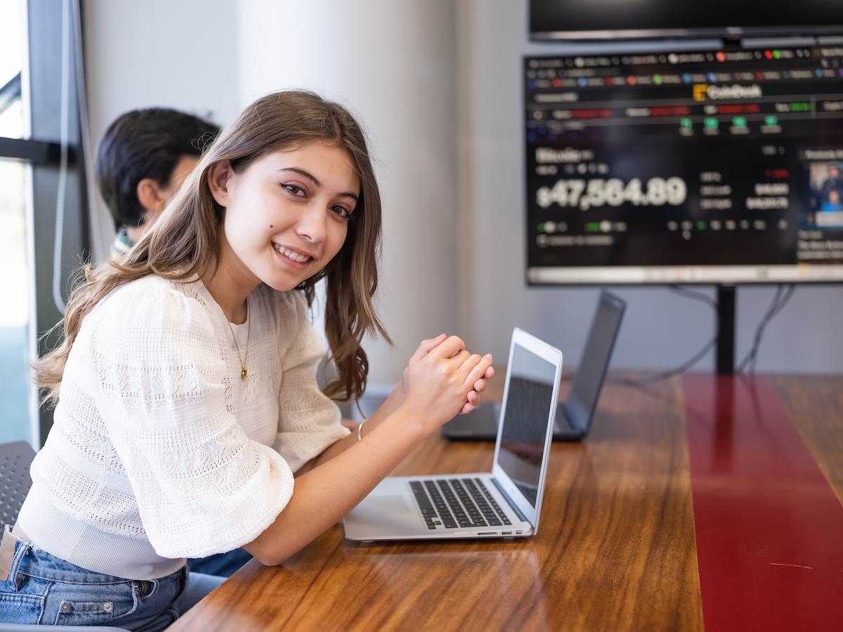 Estudiante universitaria trabajando en su laptop, con pantallas al fondo que muestran información financiera en tiempo real.