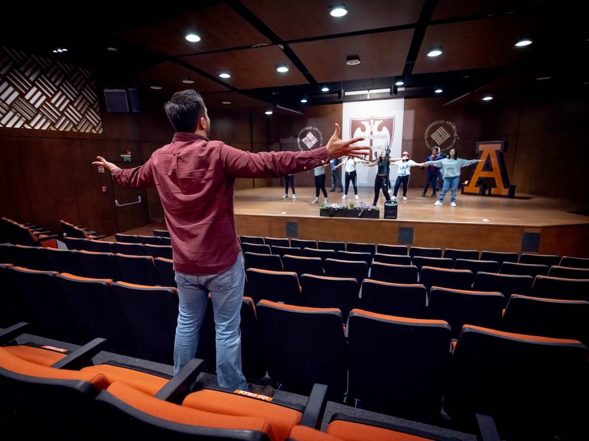 Hombre con brazos extendidos de pie entre las butacas dirigiendo un ensayo escénico, mientras un grupo de estudiantes practica una coreografía sobre el escenario de un auditorio universitario con logotipos institucionales en el fondo.