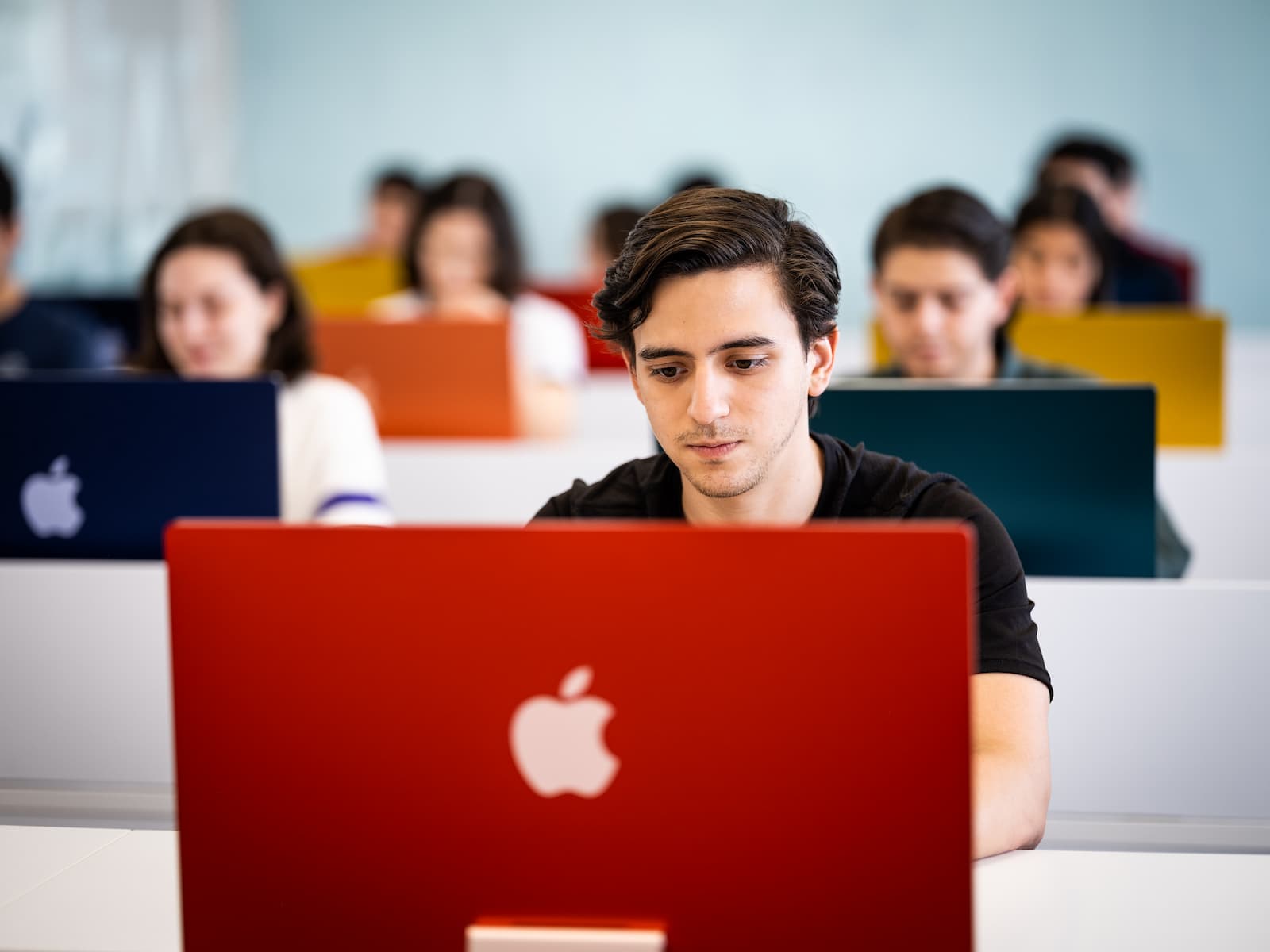 Estudiante en primer plano utilizando una iMac roja, rodeado de otros compañeros en un entorno académico luminoso y tecnológico.
