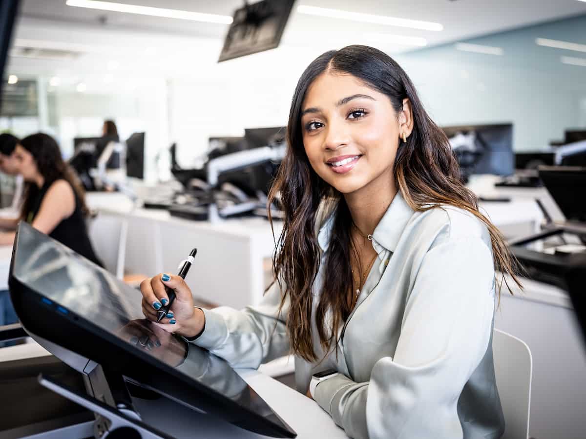 Joven creativa sonriendo mientras trabaja en una ilustración digital en aula equipada con estaciones de trabajo gráficas.