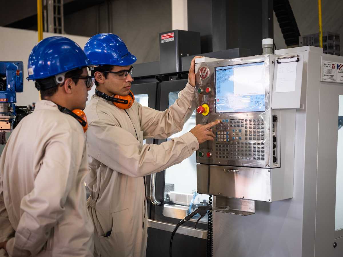 Dos estudiantes con casco azul, overol claro y audífonos protectores manipulan el panel de control de una máquina CNC en un laboratorio de manufactura avanzada.