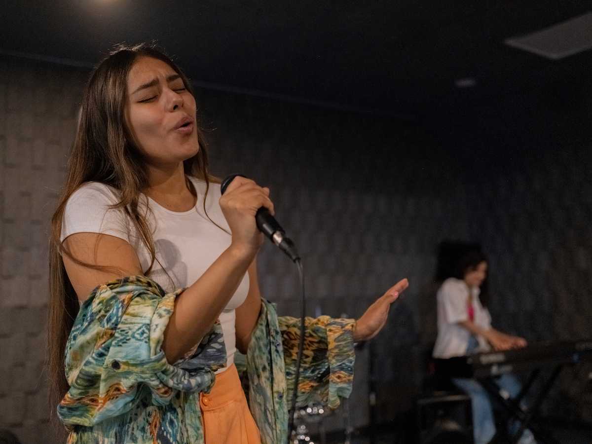 Joven cantante con camiseta blanca y kimono colorido interpretando apasionadamente una canción con micrófono en mano, mientras al fondo otra estudiante toca el teclado durante un ensayo en una sala con tratamiento acústico.