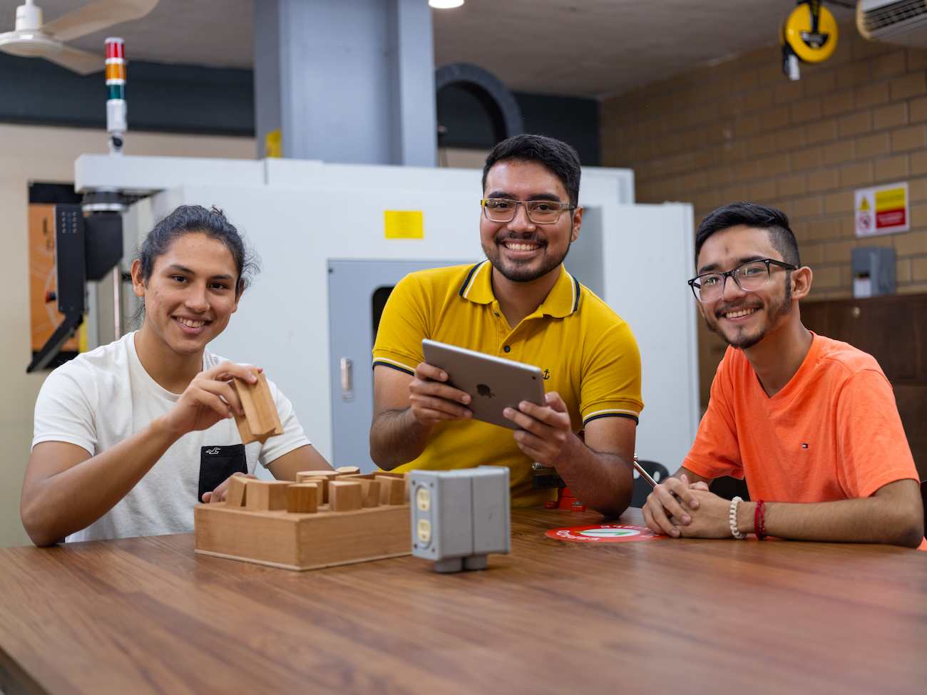 Tres estudiantes sonrientes sentados en una mesa de trabajo en un taller de diseño o manufactura; uno sostiene una pieza de madera, otro utiliza una tablet, y el tercero observa con entusiasmo, rodeados de herramientas y maquinaria industrial en el fondo.