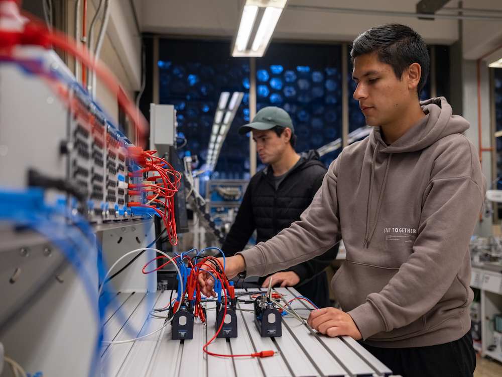 Dos estudiantes trabajando en un laboratorio de ingeniería, manipulando cables y dispositivos de prueba conectados a una mesa de experimentación electrónica, rodeados de equipos y paneles con conexiones en un entorno técnico e iluminado.