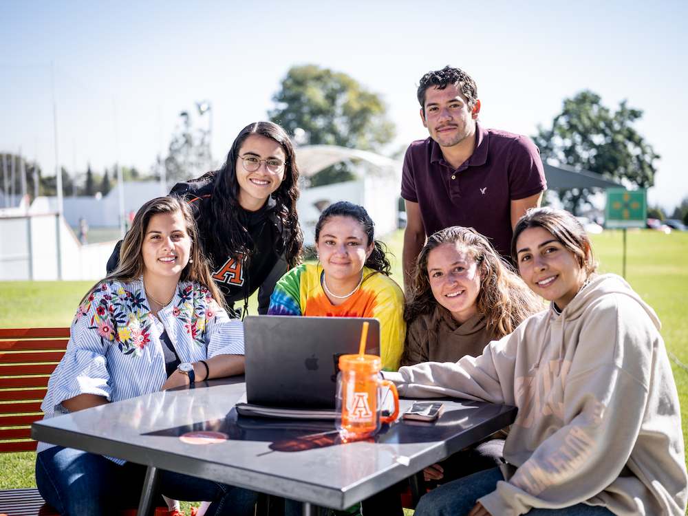 Estudiantes de la Universidad Anáhuac trabajando juntos en una laptop en una mesa al aire libre, mostrando un ambiente de colaboración y amistad.