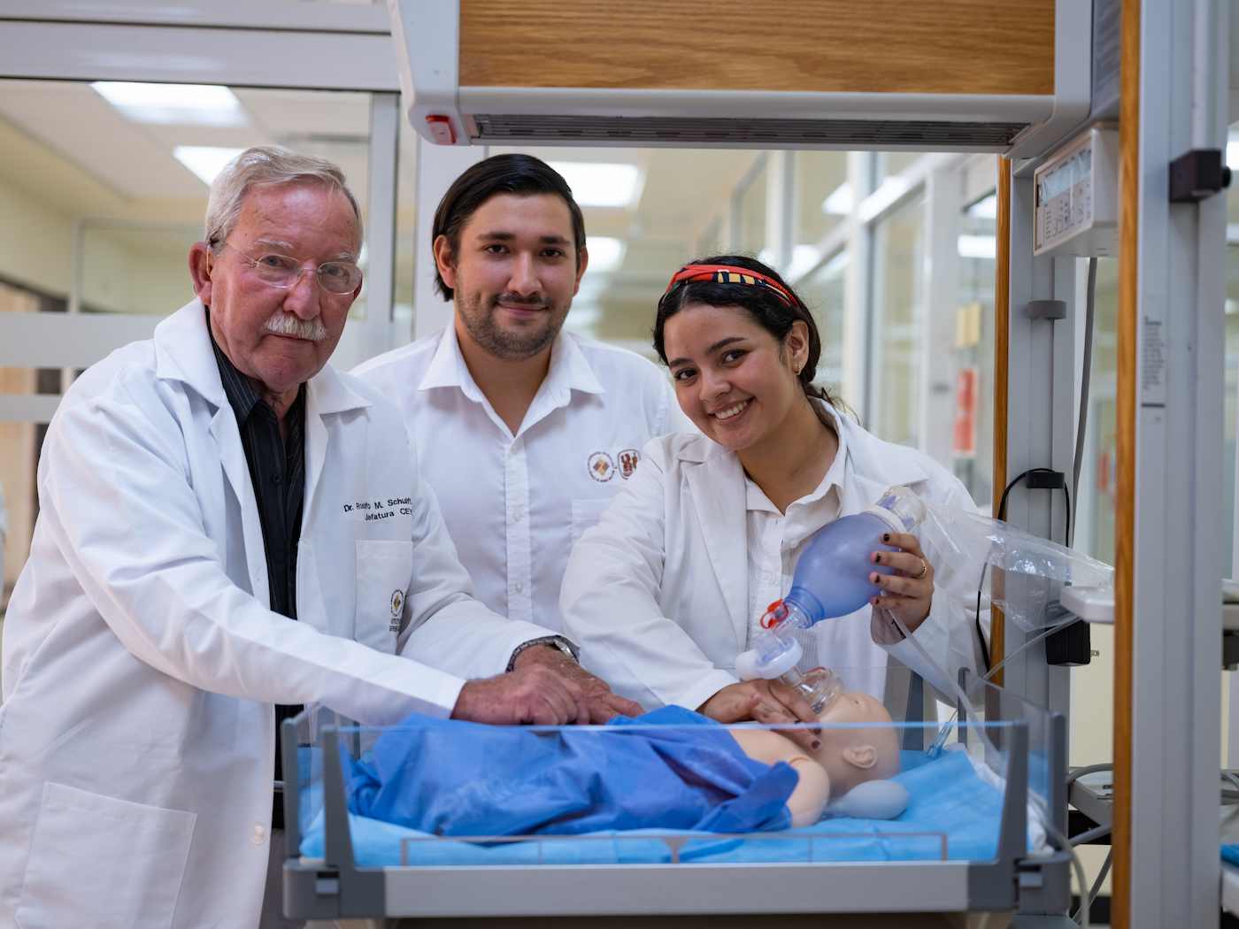 Profesor y dos estudiantes de medicina practican maniobras de reanimación con un simulador de bebé en una sala clínica.