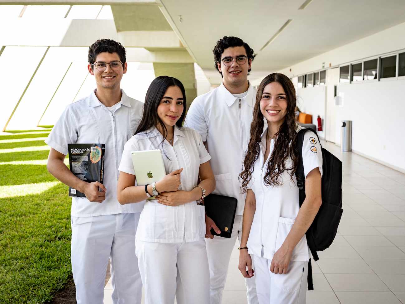 Grupo de cuatro estudiantes de medicina posan sonrientes en un pasillo universitario, vestidos con uniforme blanco y cargando libros y dispositivos.