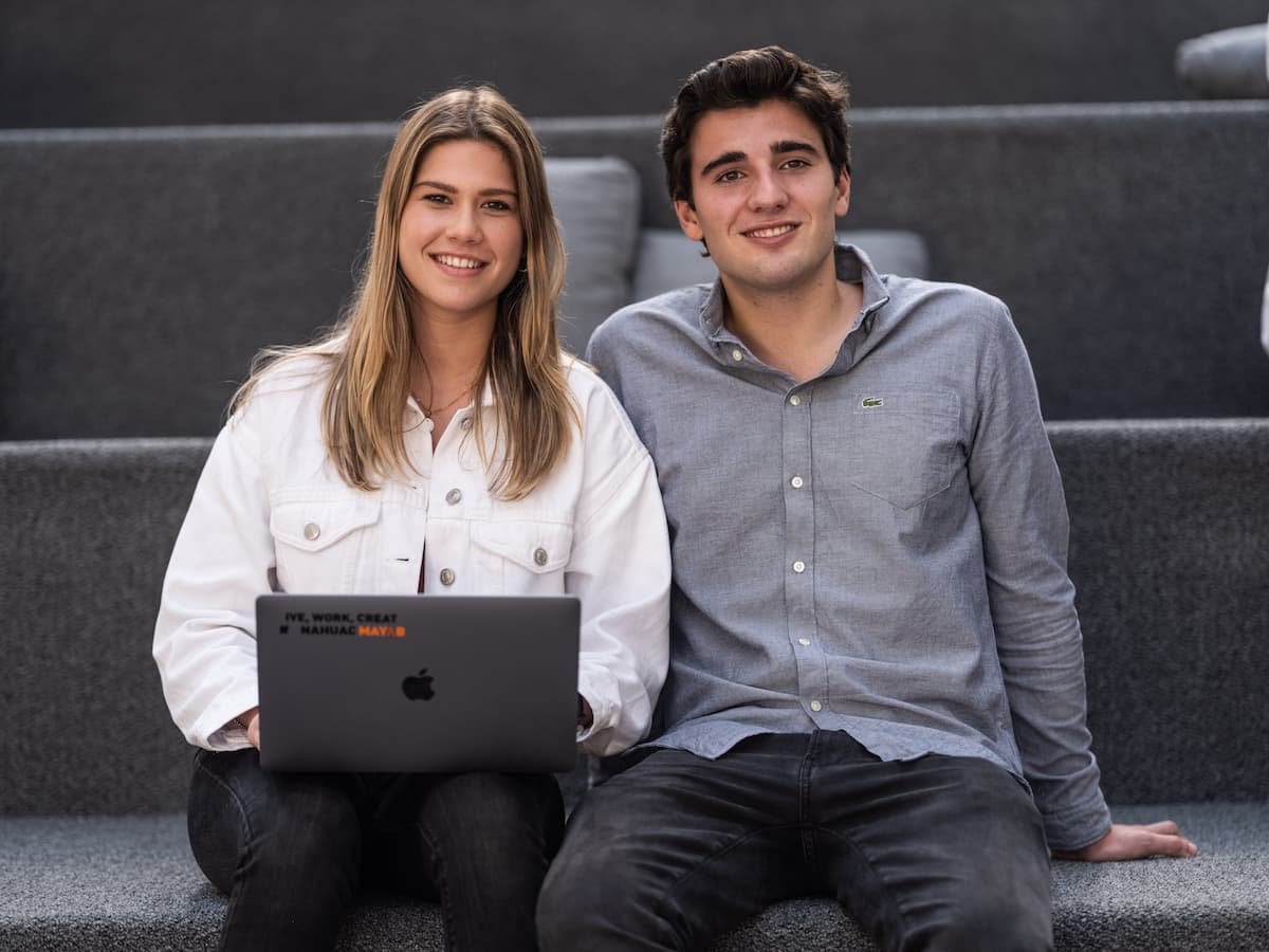 Estudiantes universitarios jóvenes, uno con laptop, sentados en gradas grises y sonriendo, reflejando un ambiente académico amigable.